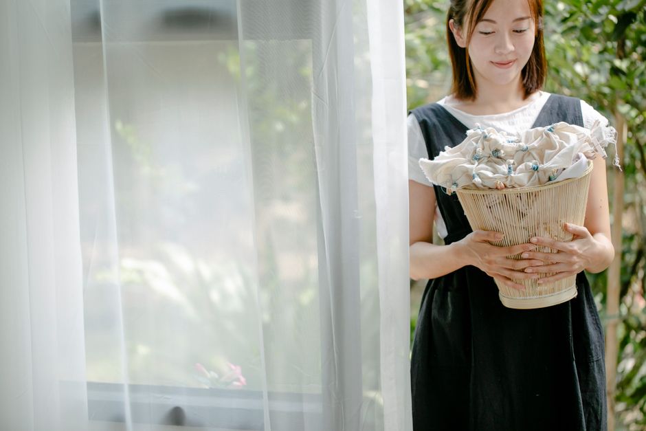 Positive Asian female in apron holding wicker basket with fabric tied with blue threads while standing near doorway on blurred background of green garden on sunny day in summer